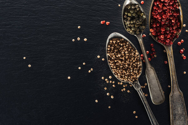 top view of pepper and coriander seeds in silver spoons on black textured background