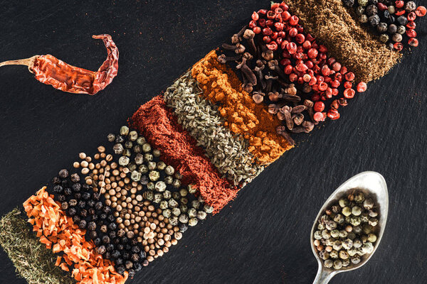 top view of traditional indian spices on textured black background near spoon with white pepper and dried chili pepper