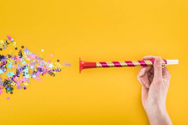 cropped view of woman holding party horn near confetti on yellow background clipart