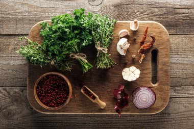 top view of green thyme and parsley on wooden chopping board near garlic cloves, red onion, dried chili peppers and pink peppercorn on wooden brown table clipart