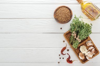 top view of black pepper, dried chili peppers, coriander in bowl near oil in bottle, board with garlic cloves and thyme on white wooden table clipart