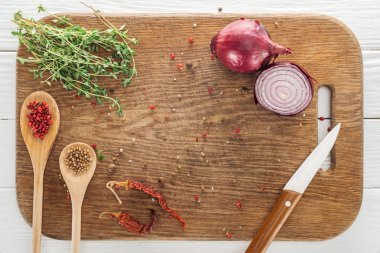 top view of thyme, knife, spoons with coriander and pink peppercorn, dried chili peppers and red onion on wooden chopping board clipart