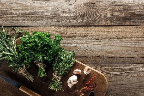 ニンニククローブ ナイフ 乾燥唐辛子 木製茶色のテーブルの近くの木製まな板上の緑のタイム パセリ ローズマリーのトップビュー — ストック写真