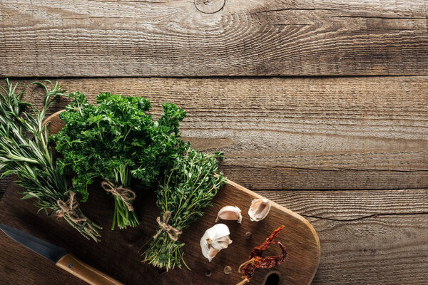 top view of green thyme, parsley, rosemary on wooden chopping board near garlic cloves, knife, dried chili pepper and black pepper on wooden brown table