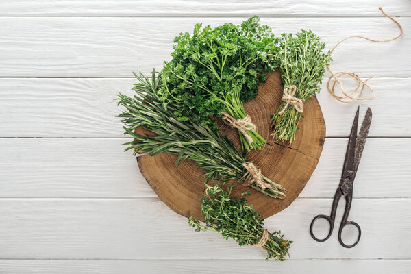 top view of green parsley, rosemary and thyme on brown stump near vintage scissors on white wooden table