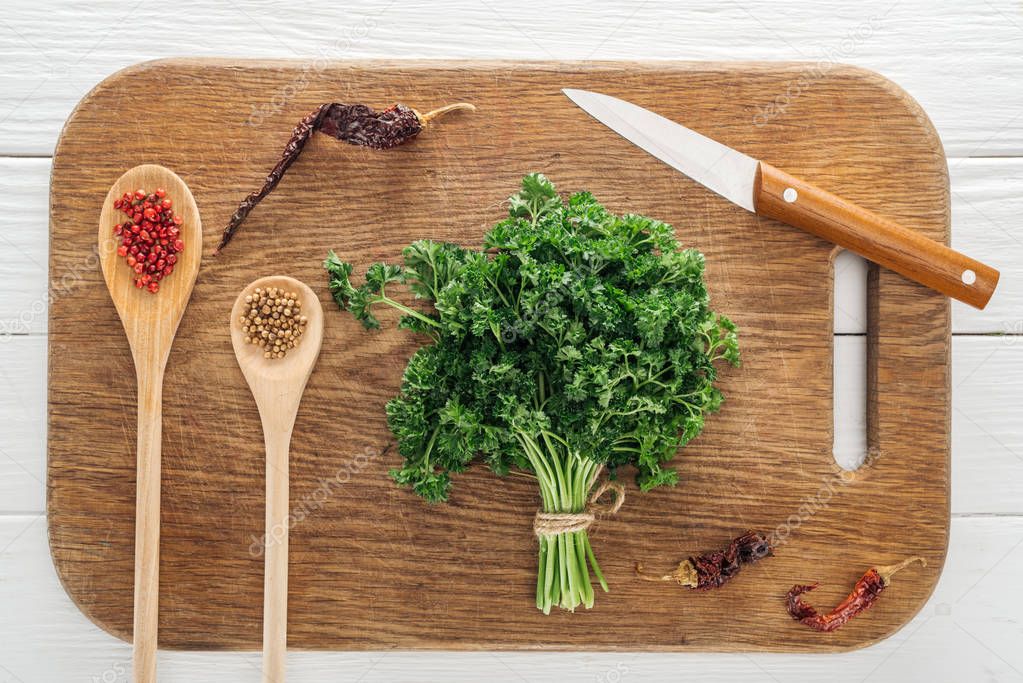 top view of green parsley, knife, spoons with coriander and pink peppercorn, dried chili peppers on wooden chopping board