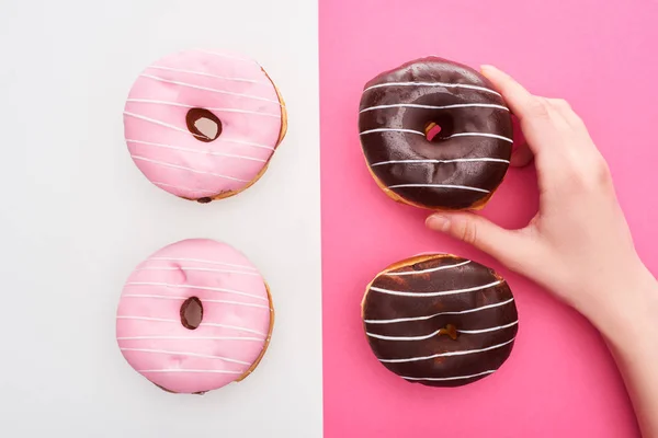 在白色和粉红色背景上接近巧克力和粉红色甜甜圈的雌性手的裁剪视图 — 图库照片