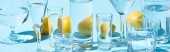 panoramic shot of transparent glasses with water and whole lemons on blue background 