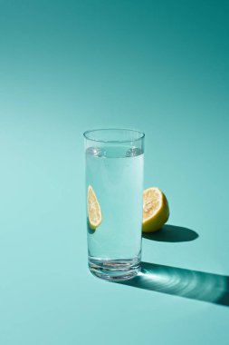 transparent glass with fresh water and lemon half on turquoise background clipart