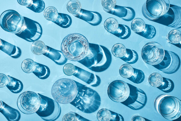 вид сверху на стаканы с прозрачной водой и тени на голубой поверхности

