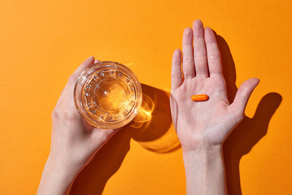 обрезанный вид женщины, держащей таблетку на ладони возле стекла с водой на оранжевом фоне
