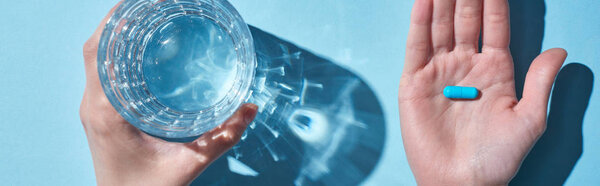 обрезанный вид женщины, держащей голубую таблетку на ладони и стекло с водой на синем фоне, панорамный снимок
