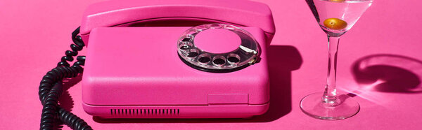 panoramic shot of transparent glass with cocktail and olive near vintage dial phone on pink background