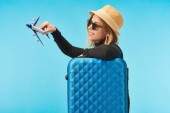 Blondes glückliches Mädchen mit Sonnenbrille und Strohhut mit Spielzeugflugzeug nahe blauer Reisetasche isoliert auf blauem Grund
