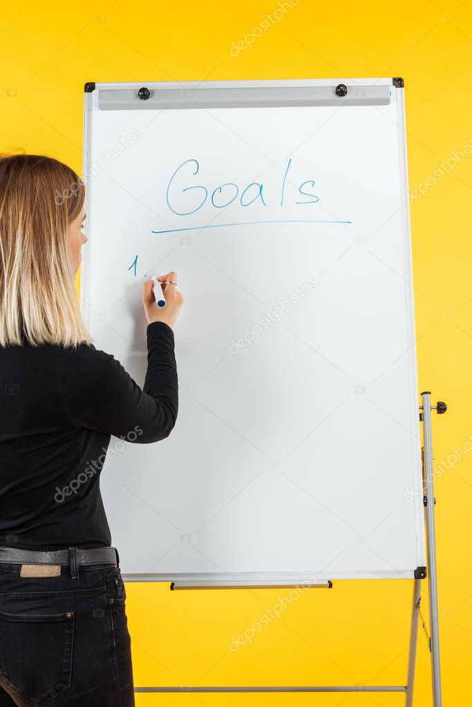 back view of businesswoman standing near white flipchart, writing goals list