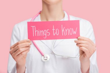 cropped view of doctor in white coat and stethoscope holding card with things to know lettering isolated on pink clipart