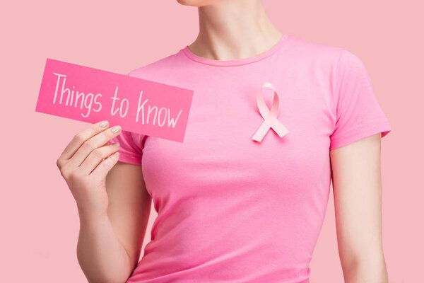 cropped view of woman in pink t-shirt holding card with things to know lettering isolated on pink, breast cancer concept