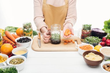 cropped view of woman in apron adding sliced carrot at jar with salad on wooden table isolated on white clipart