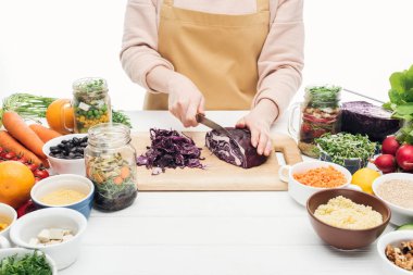 cropped view of woman in apron cutting red cabbage on wooden table isolated on white clipart