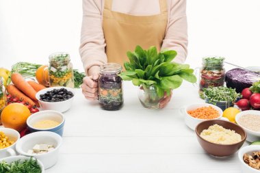beyaz izole ahşap masa üzerinde salata ve yeşil yaprakları ile cam kavanoz tutan önlük kadın kırpılmış görünümü
