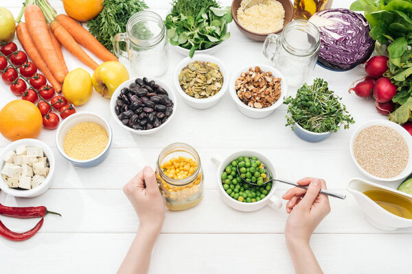 cropped view of woman adding peas in jar with salad on wooden white table 