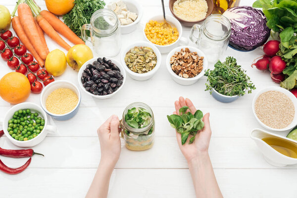 cropped view of woman adding herbs in glass jar with salad on wooden white table 