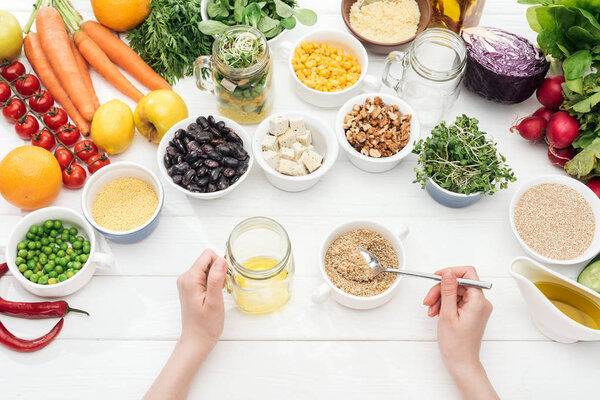 cropped view of woman adding seeds in jar with salad on wooden white table 