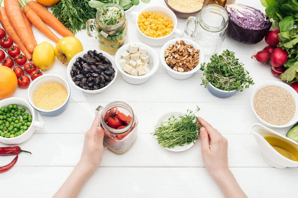 cropped view of woman holding glass jar with salad and plate with herbs on wooden white table