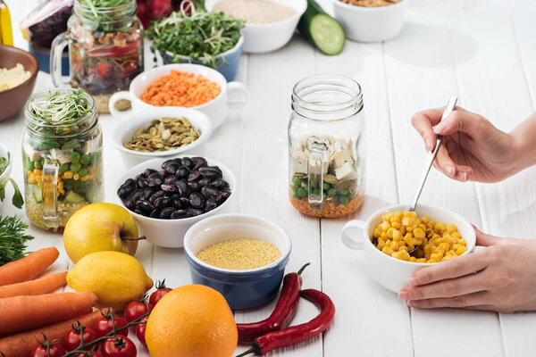 cropped view of woman adding corn in glass jar on wooden white table