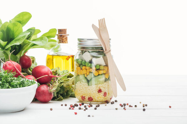 fresh vegetable salad in glass jar near oil, radish and spices isolated on white