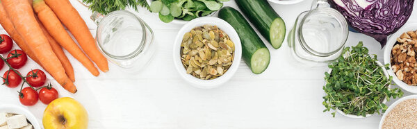 top view of tasty fruits and vegetables in bowls near glass jars on wooden white table with copy space, panoramic shot
