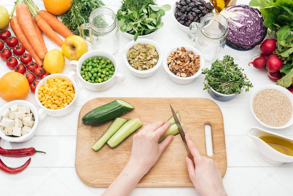 cropped view of woman cutting cucumber on wooden white table