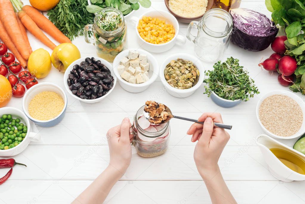 cropped view of woman adding nuts in jar with salad on wooden white table 