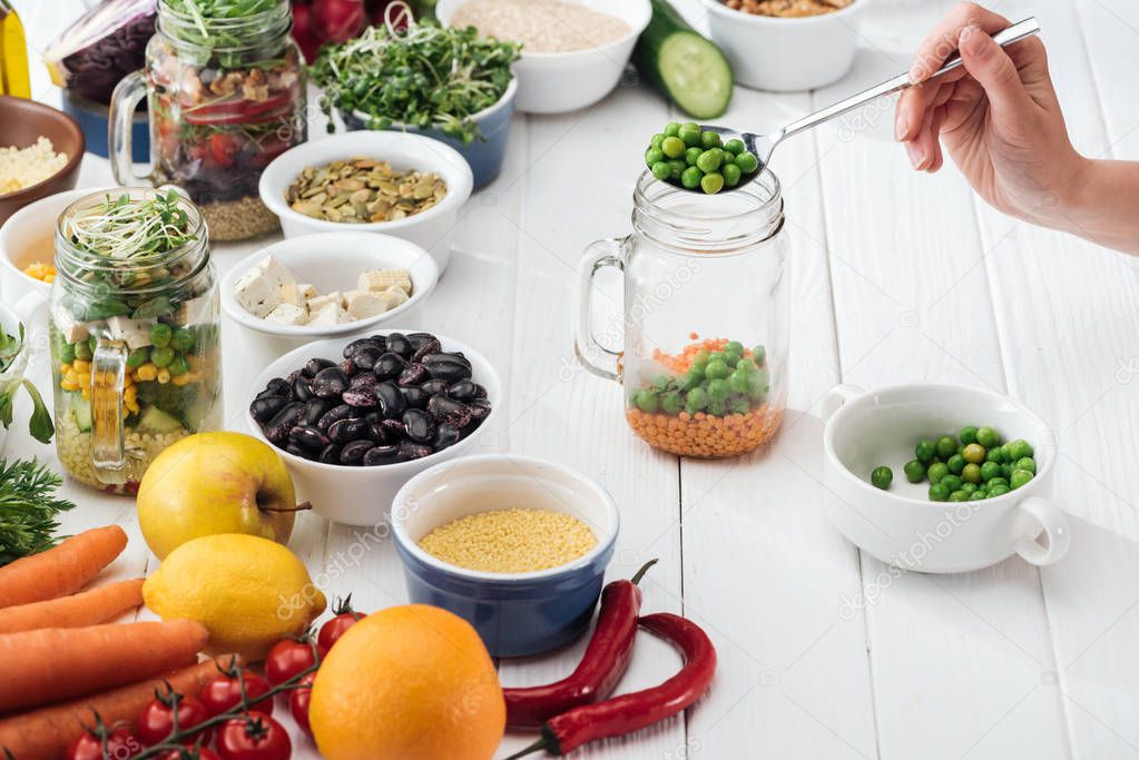 cropped view of woman adding green peas in glass jar on wooden white table