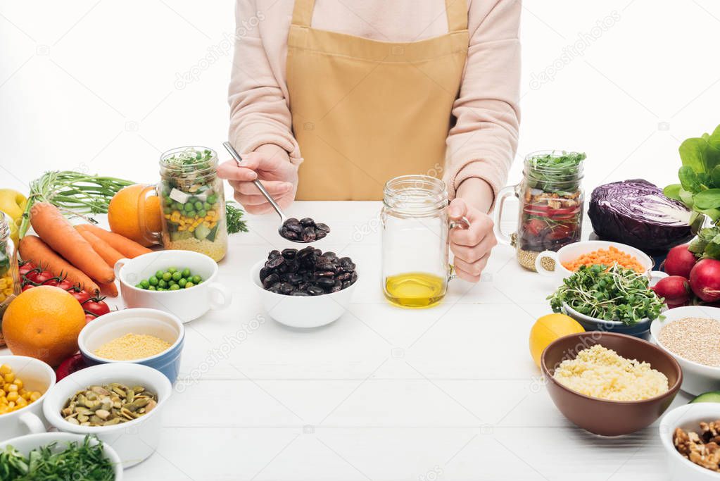 cropped view of woman in apron adding beans at jar with oil on wooden table isolated on white