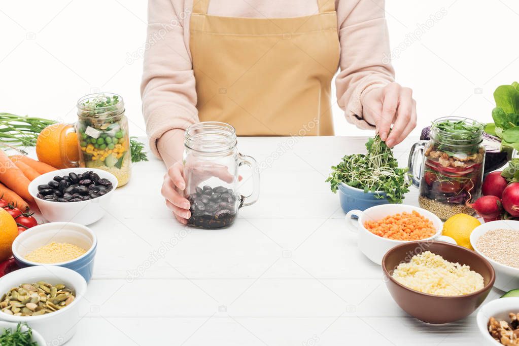 cropped view of woman in apron adding greens to glass jar with beans on wooden table isolated on white