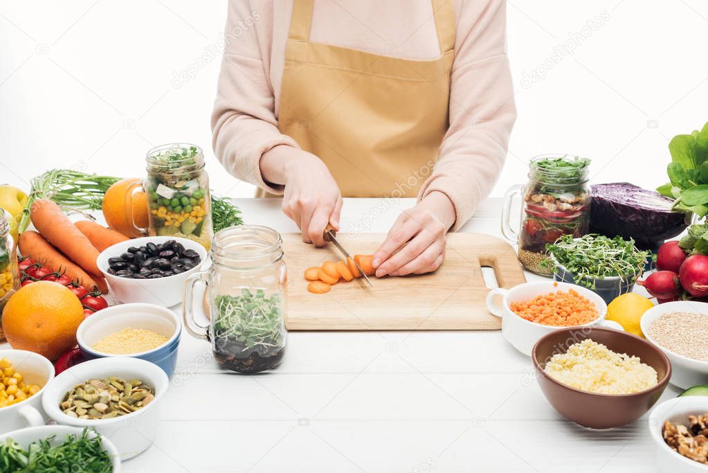 cropped view of woman in apron cutting carrots on wooden table isolated on white