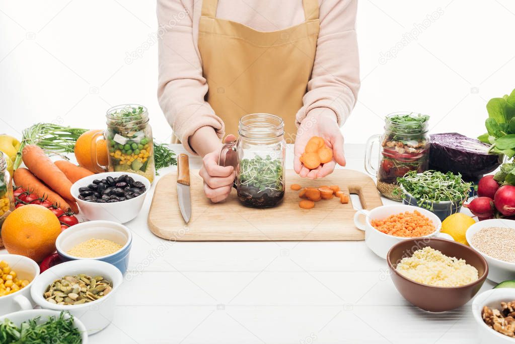 cropped view of woman in apron adding sliced carrot at jar with salad on wooden table isolated on white