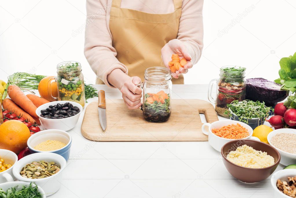 partial view of woman in apron adding sliced carrot at jar with salad on wooden table isolated on white