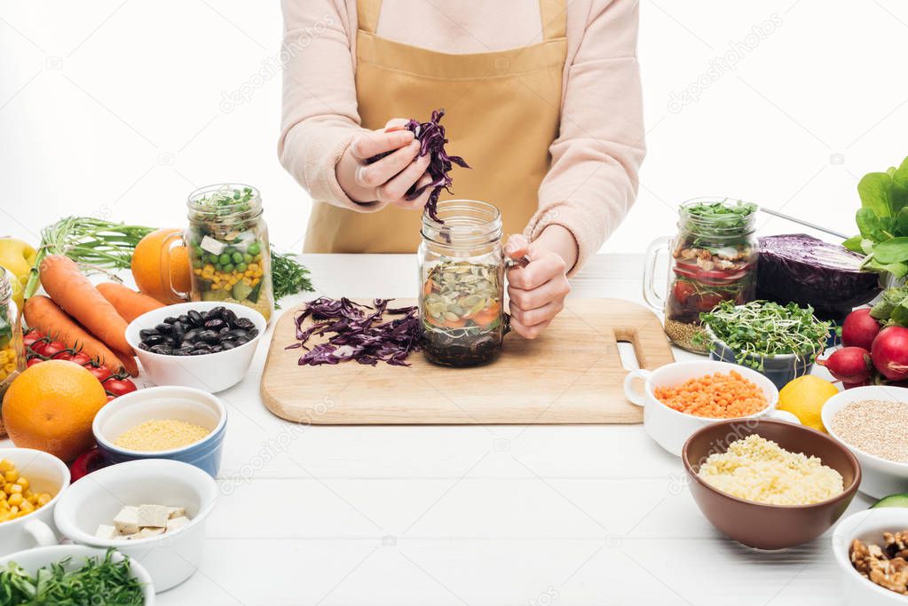 cropped view of woman in apron adding sliced red cabbage in glass jar on wooden table isolated on white