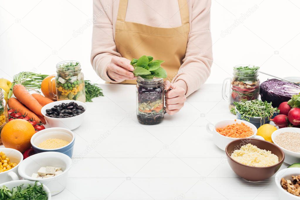 cropped view of woman in apron adding green leaves in glass jar on wooden table isolated on white