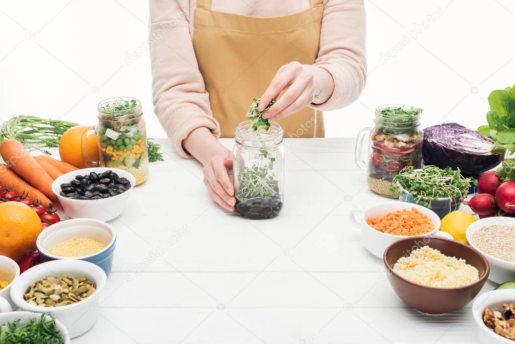 cropped view of woman in apron adding sprouts in glass jar on wooden table isolated on white