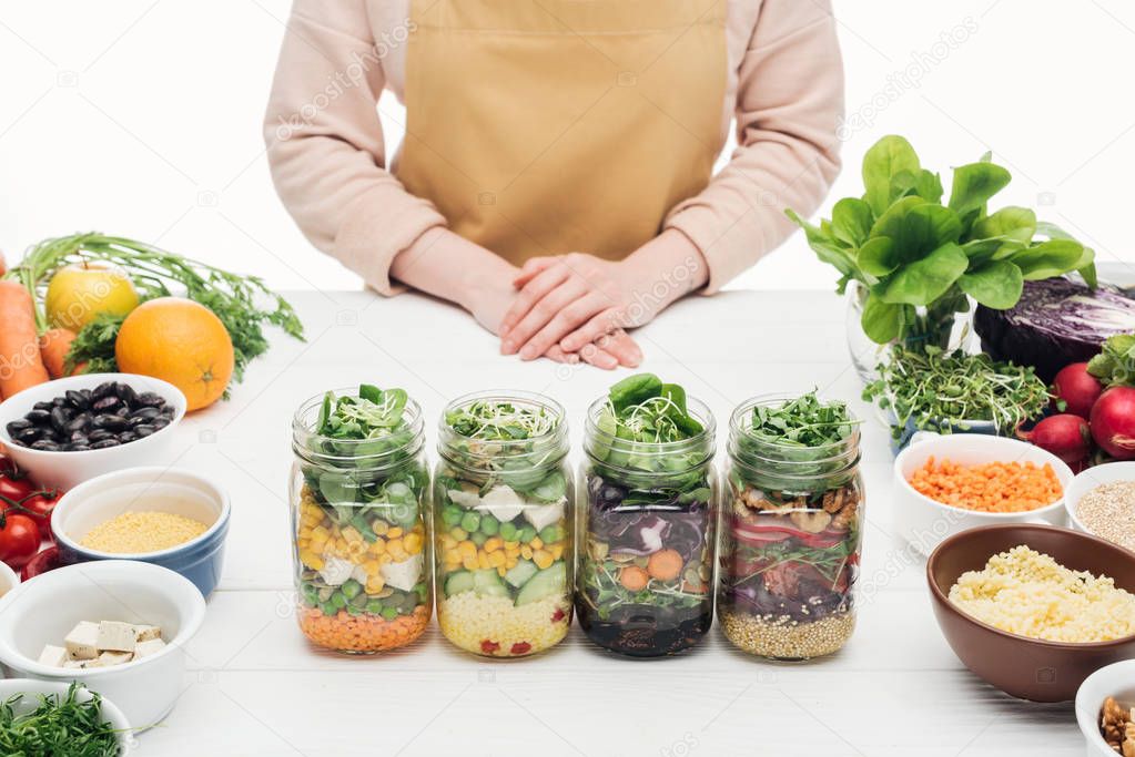 partial view of woman in apron standing near glass jars with salad on wooden table isolated on white