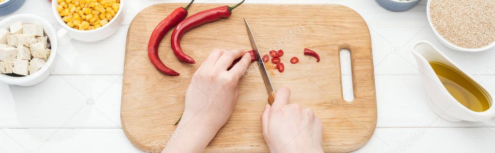 cropped view of woman cutting chili peppers on wooden chopping board on  white table, panoramic shot