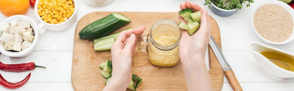 cropped view of woman adding chopped cucumber to couscous in glass jar on wooden chopping board, panoramic shot