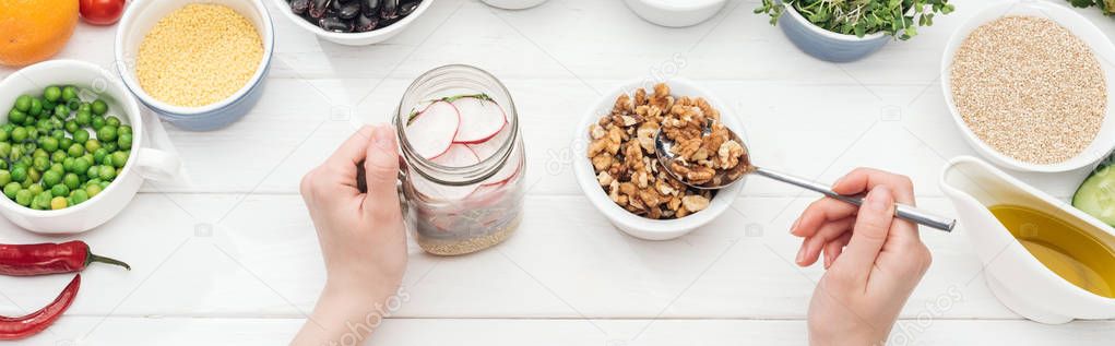 cropped view of woman adding nuts in jar with salad on wooden white table, panoramic shot