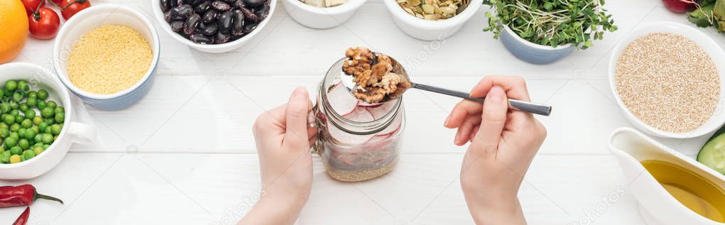 cropped view of woman adding nuts in glass jar with salad on wooden white table, panoramic shot