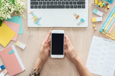 dizüstü bilgisayar, haftalık liste, kırtasiye ve elinde akıllı telefon tutan ahşap masa arkasında oturan kadın kırpılmış görünümü
