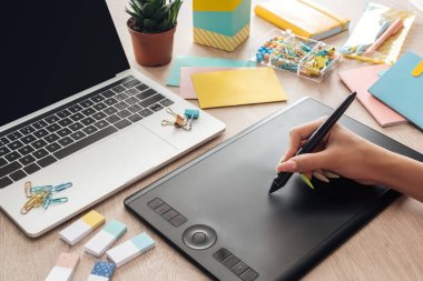 cropped view of woman working with drawing tablet, sitting behind wooden table with stationery and laptop clipart