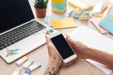 dizüstü bilgisayar ve kırtasiye ile ahşap masa arkasında oturan kadın kırpılmış görünümü, elinde akıllı telefon tutarak
